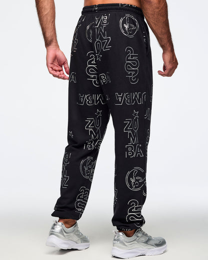 Black jogger sweatpants shown from the back with an allover silver Zumba logo, icon, and star print and cuffed ankles. Male model is wearing the relaxed-fit joggers with silver athletic sneakers.