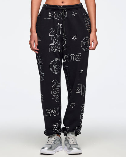 Black jogger sweatpants with an allover silver Zumba logo, icon, and star print, elastic drawstring waistband, and cuffed ankles. Female model is wearing the relaxed-fit joggers with silver athletic sneakers.