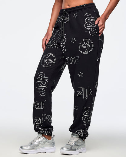 Black jogger sweatpants with an allover silver Zumba logo, icon, and star print, elastic drawstring waistband, and cuffed ankles. Female model is wearing the relaxed-fit joggers with silver athletic sneakers in a side view.