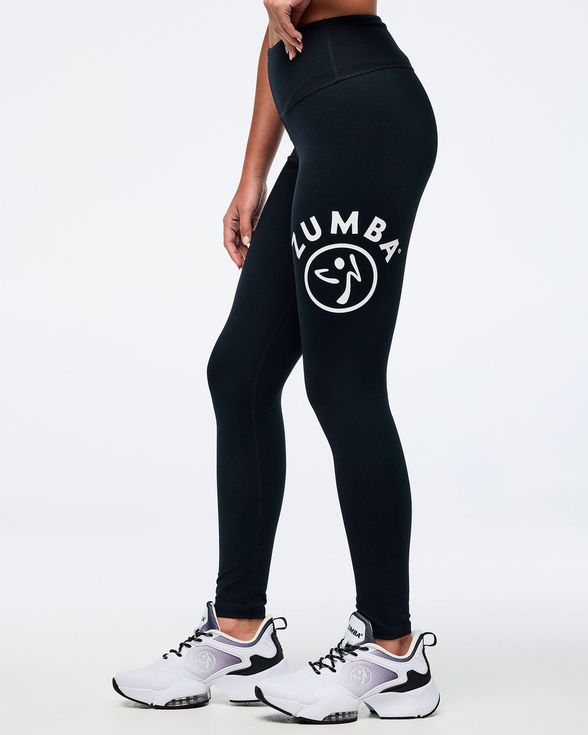 A female model wearing black high-waisted Zumba leggings with a white Zumbito logo on the left thigh, paired with white and black athletic sneakers, shown from a side profile.