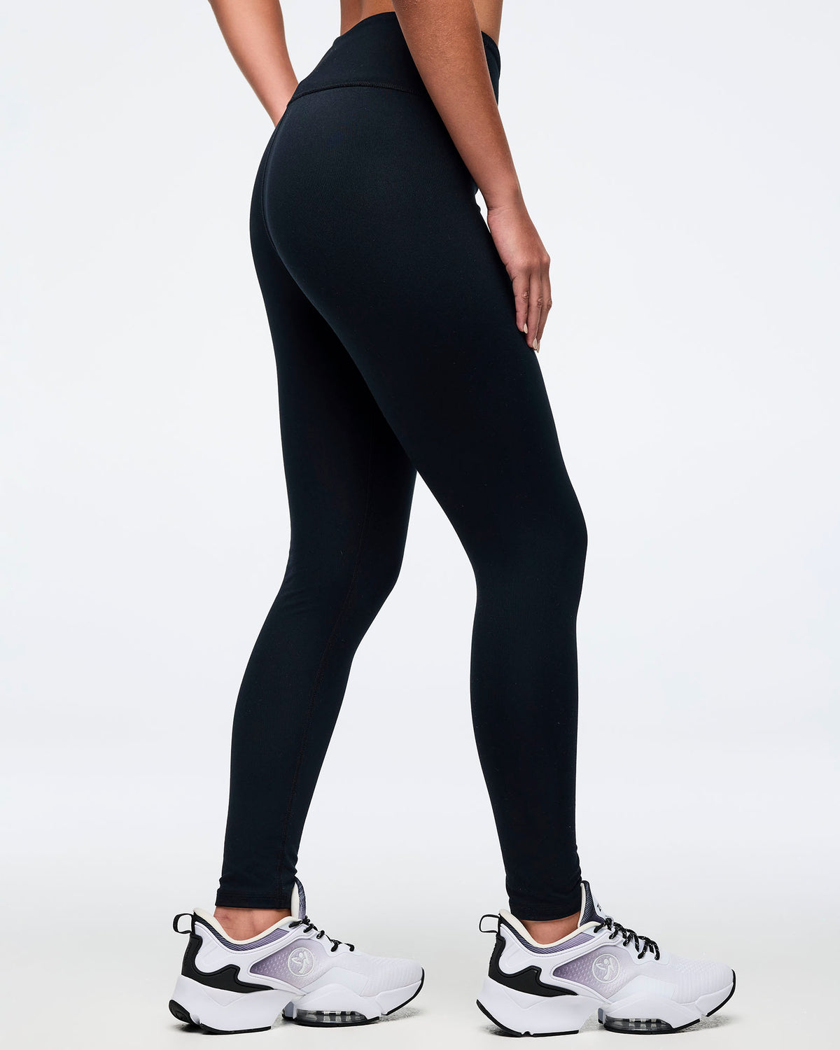 Back view of a female model wearing black high-waisted Zumba leggings, paired with white and black athletic sneakers.
