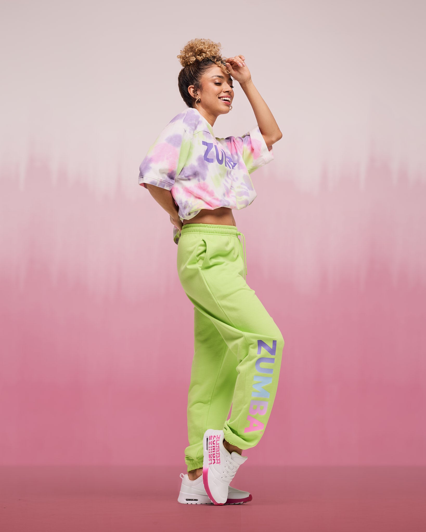 Female model wearing a cropped pastel tie-dye patterned, short-sleeve top with “ZUMBA” lettering, paired with lime green jogger sweatpants featuring large vertical “ZUMBA” text on the leg and white sneakers with pink details.