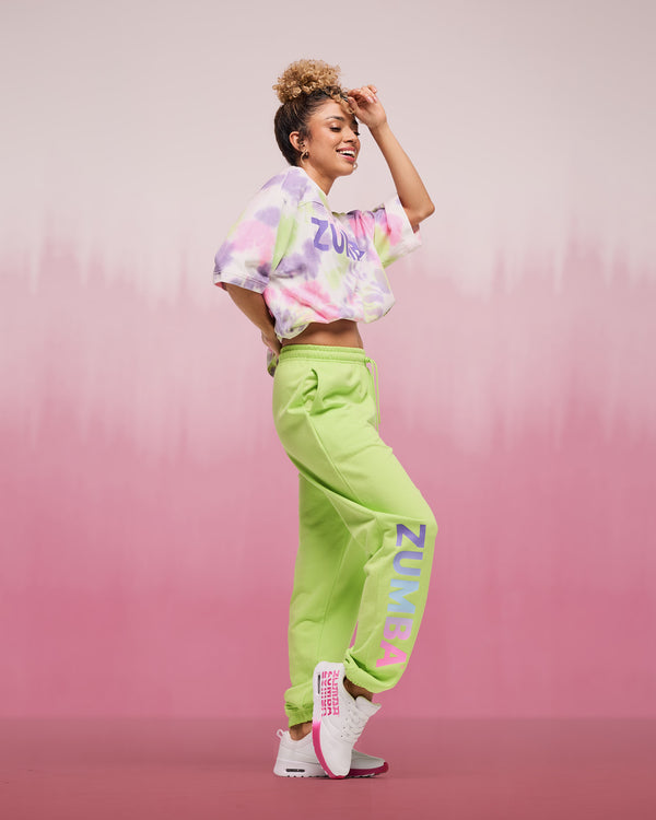 Female model wearing a cropped pastel tie-dye patterned, short-sleeve top with “ZUMBA” lettering, paired with lime green jogger sweatpants featuring large vertical “ZUMBA” text on the leg and white sneakers with pink details.