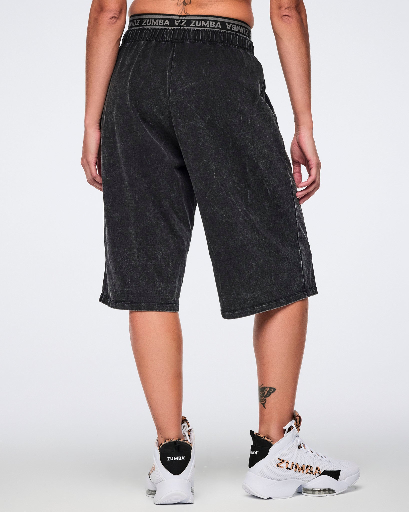 Black long pants with an oversized fit, elastic Zumba logo waistband, and below-the-knee length, shown from the back on a female model wearing white high-top Zumba sneakers with black and cheetah-print details.