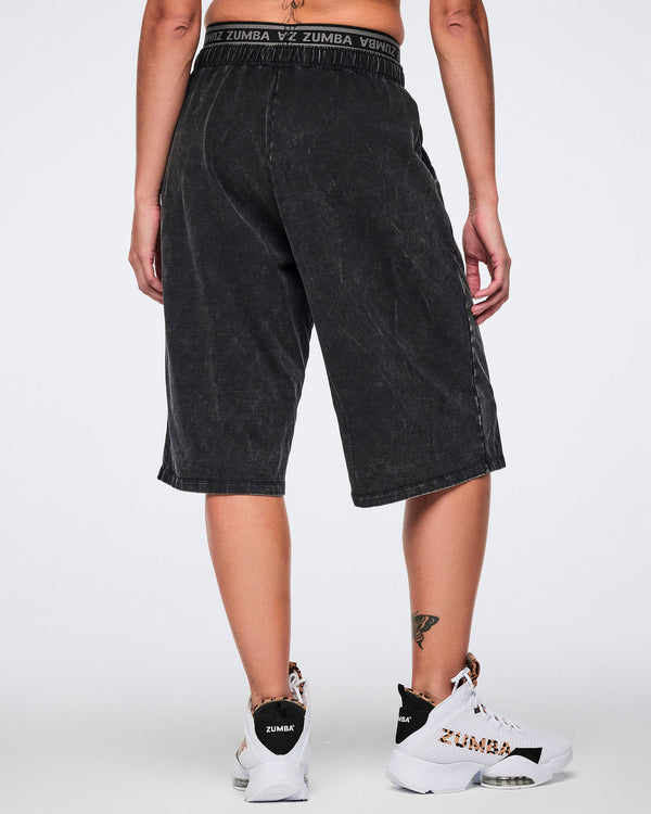 Black long pants with an oversized fit, elastic Zumba logo waistband, and below-the-knee length, shown from the back on a female model wearing white high-top Zumba sneakers with black and cheetah-print details.