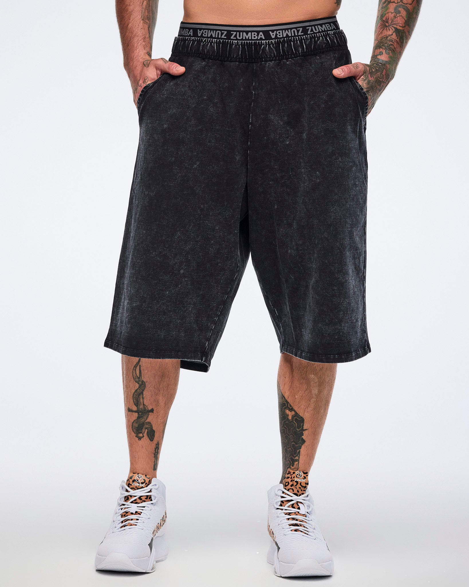 Black long pants with an oversized fit, side pockets, elastic Zumba logo waistband, and below-the-knee length, shown from the front on a male model wearing white high-top Zumba sneakers with black and cheetah-print details.