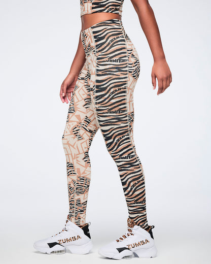 Side view of beige and black mixed-print high-waisted leggings with a fitted full-length silhouette, combining zebra stripes and abstract geometric panels. Female model wears the leggings with white high-top Zumba sneakers and a matching beige and black crop top.