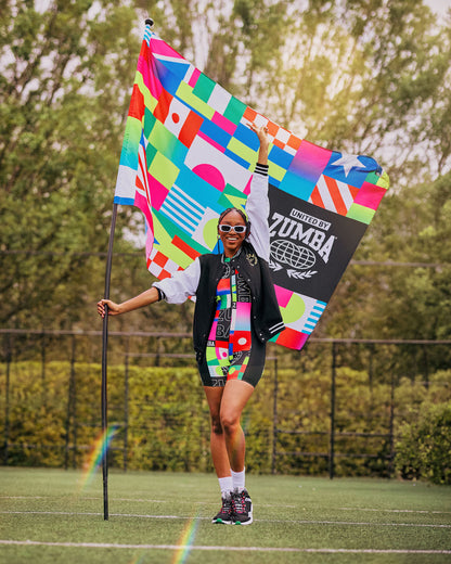 Female model outdoors posing on grass holding a patterned flag with one square in black and white with “United by Zumba” text.