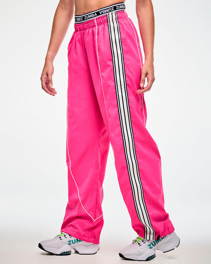 Female model wearing pink track pants with a black and white striped side panel and a 'ZUMBA' waistband, paired with white sneakers.