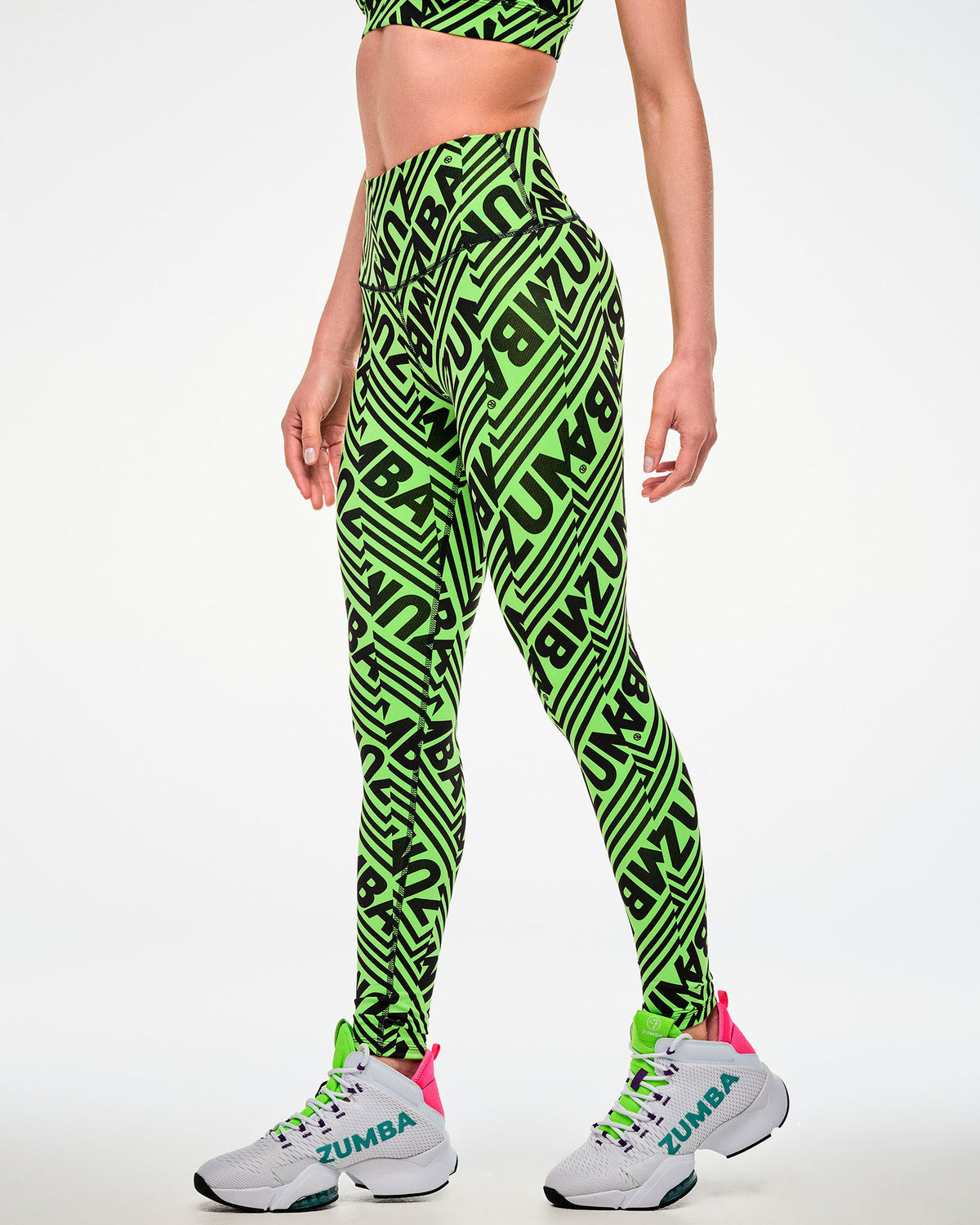 Female model wearing green leggings with a repeated black "ZUMBA" text print and paired with white shoes, side view.