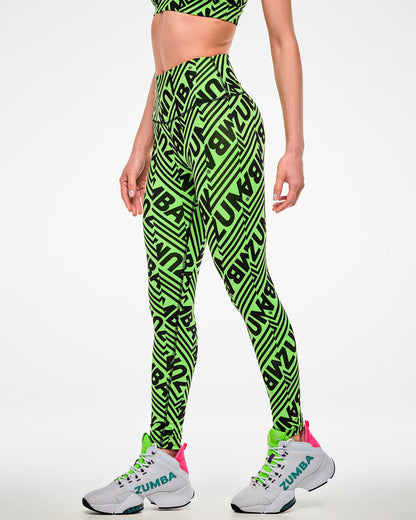 Female model wearing green leggings with a repeated black "ZUMBA" text print and paired with white shoes, side view.
