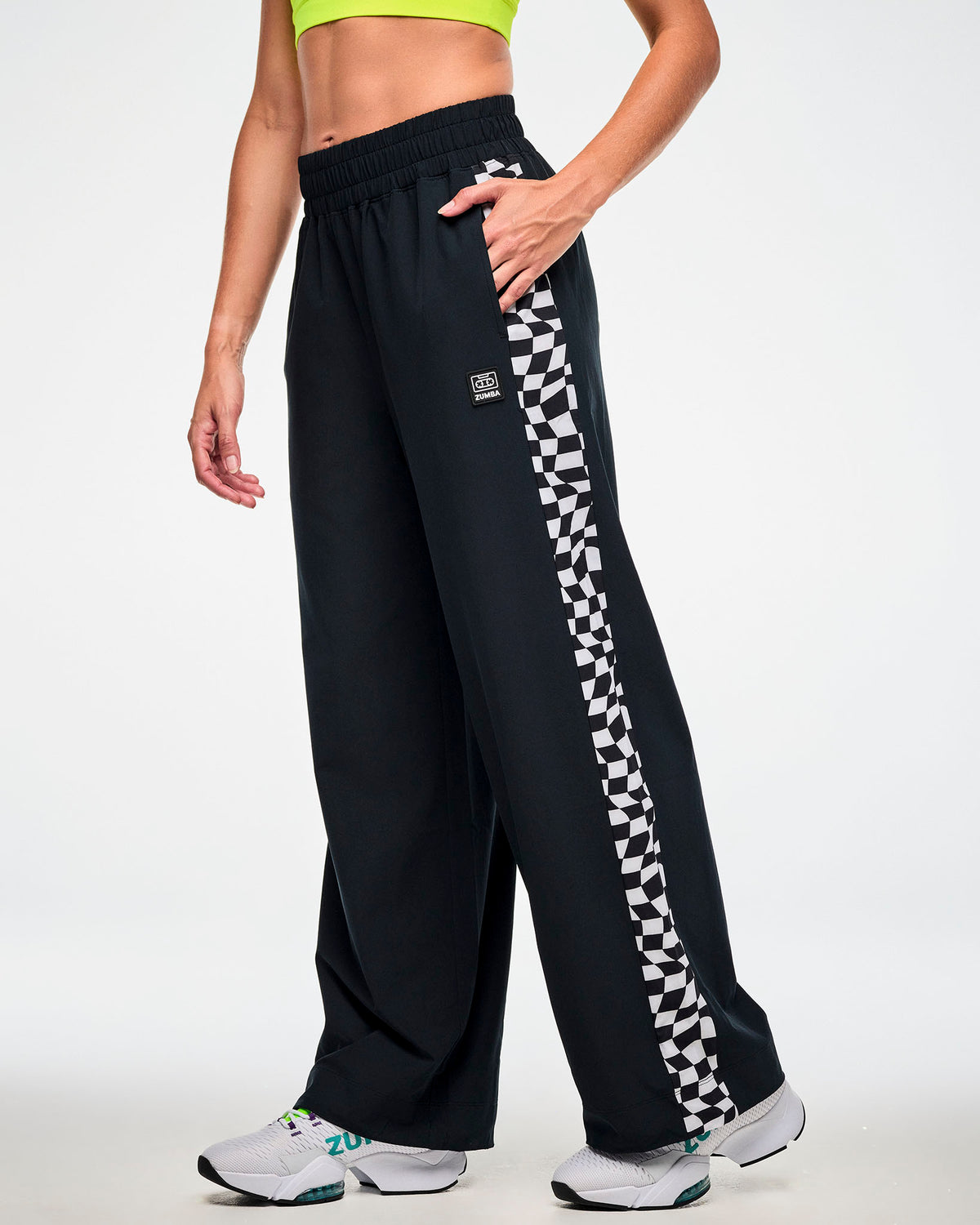 Female model wearing black track pants with a black and white checkerboard side panel and white sneakers, hand in the pocket.