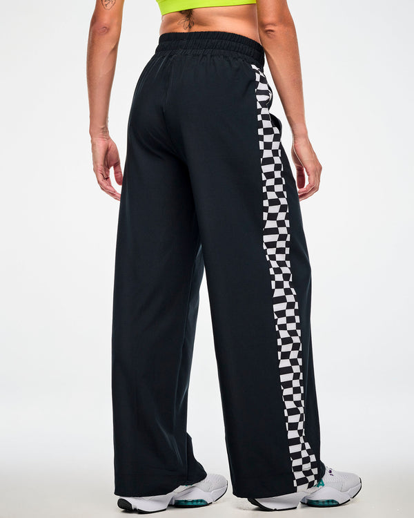 Back view of a female model wearing black track pants with a checkerboard side panel and white sneakers.