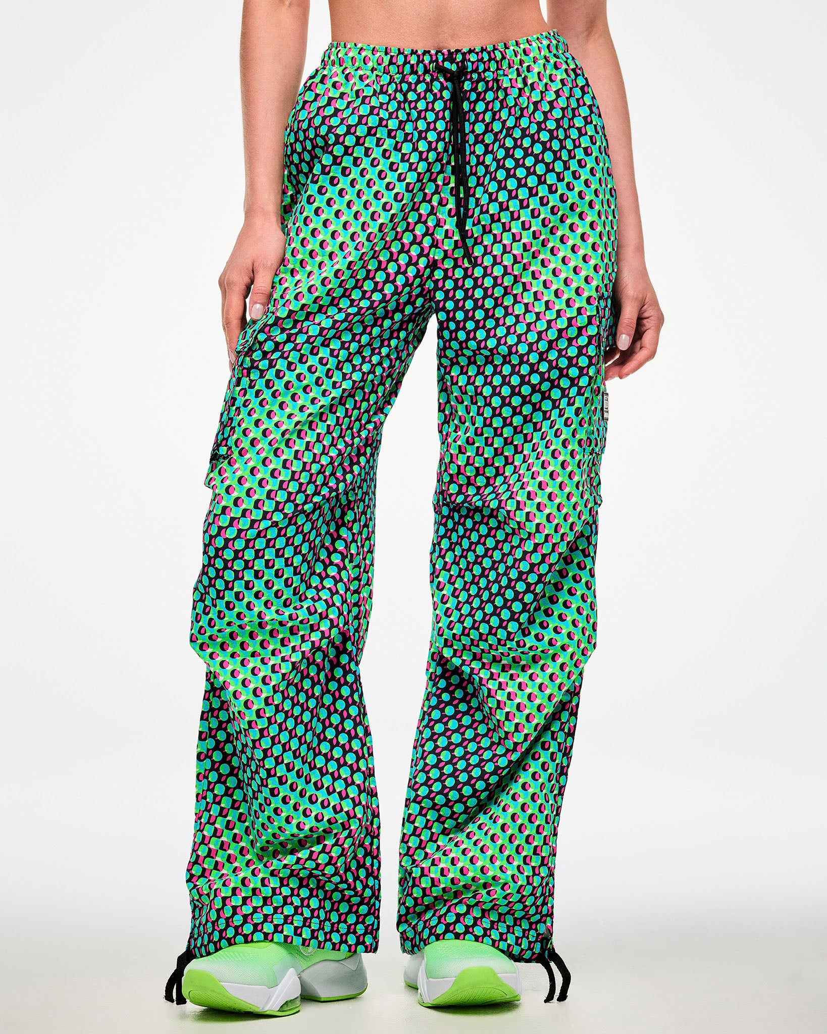 Female model wearing green cargo pants with black drawcords hanging over the aqua, black, and pink dot print, paired with green sneakers.