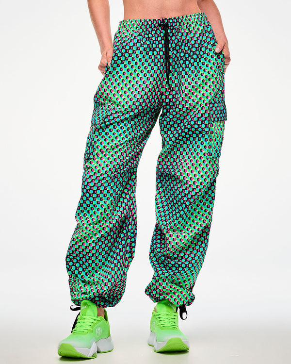 Female model wearing green cargo pants with an aqua, black, and pink dot print and green sneakers, hand in side pocket.