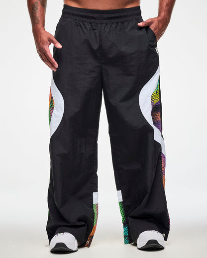Male model wearing black track pants with a white and multicolor dot-print side panel and white sneakers, one hand in the side pockets.