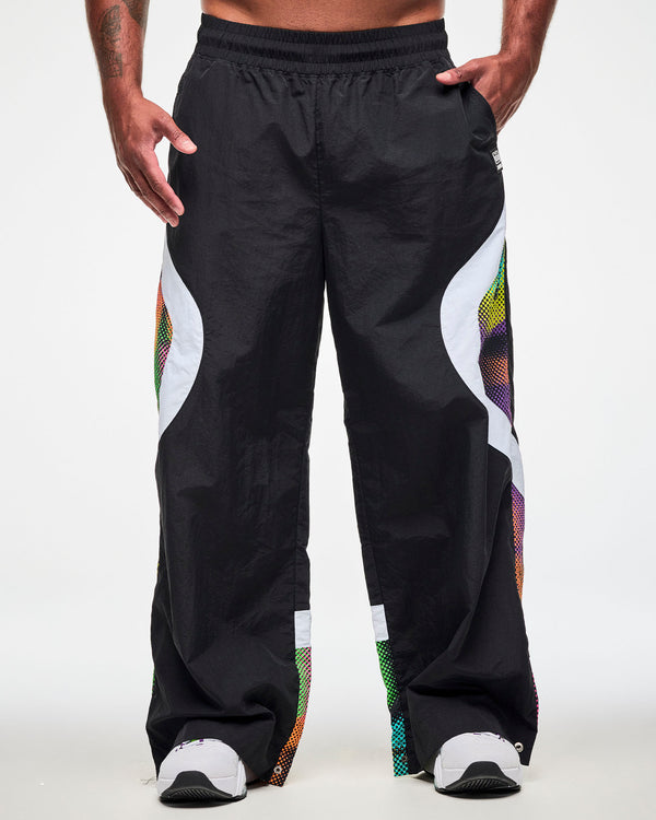 Male model wearing black track pants with a white and multicolor dot-print side panel and white sneakers, one hand in the side pockets.