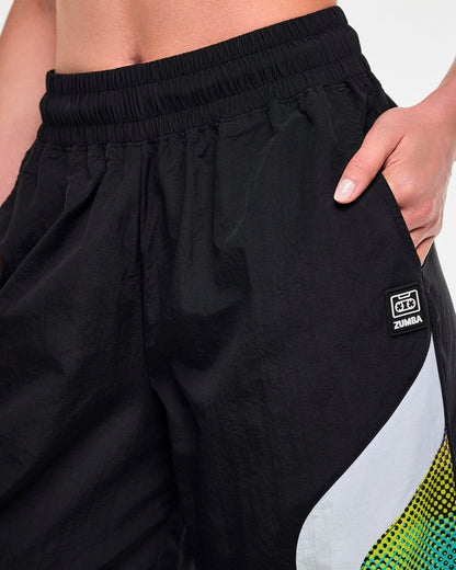 Close-up of a female model with a hand in the left pocket of black track pants, showing a black tag featuring a cassette tape icon and "ZUMBA" text, both in white, in more detail.