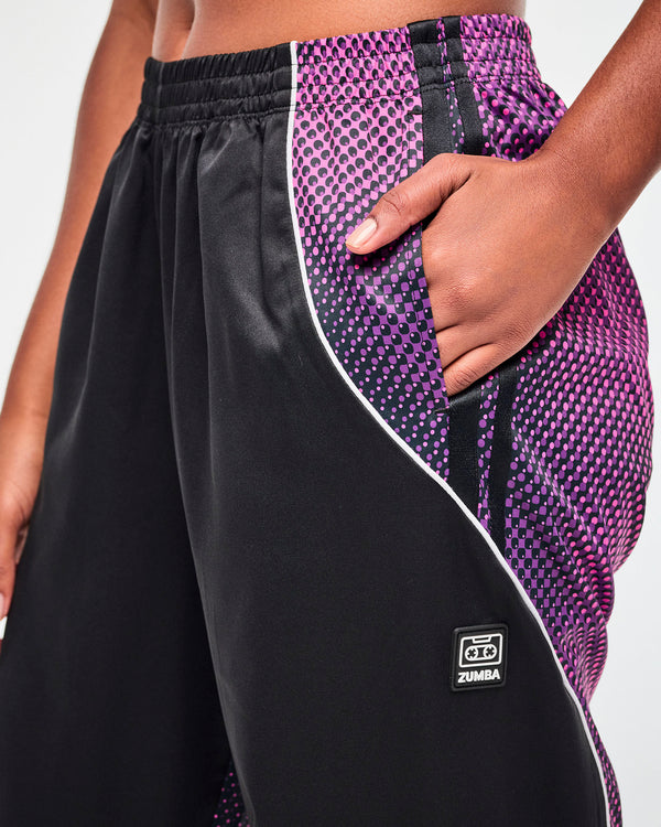 Close-up of the left side pocket on black track pants, where the purple-to-pink gradient panel meets the black front, with a black tag featuring a cassette tape icon and 
