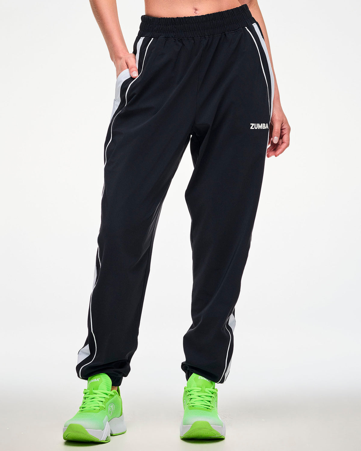Female model wearing black joggers with a white side stripe, "ZUMBA" text, and green shoes, hand in the side pocket.