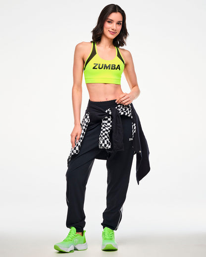 Female model wearing a yellow sports bra with black joggers and green shoes, a checkered shirt tied at the waist.