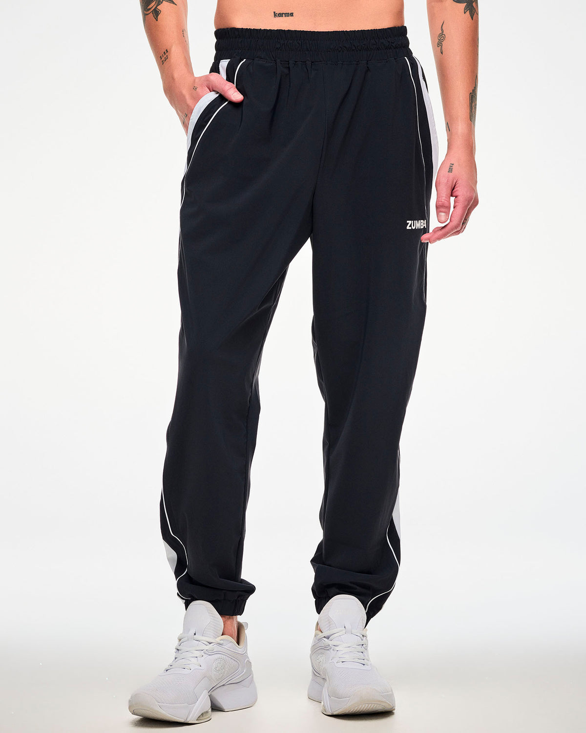 Male model wearing black joggers with a white side stripe, "ZUMBA" text, and white shoes, hand in the side pocket.