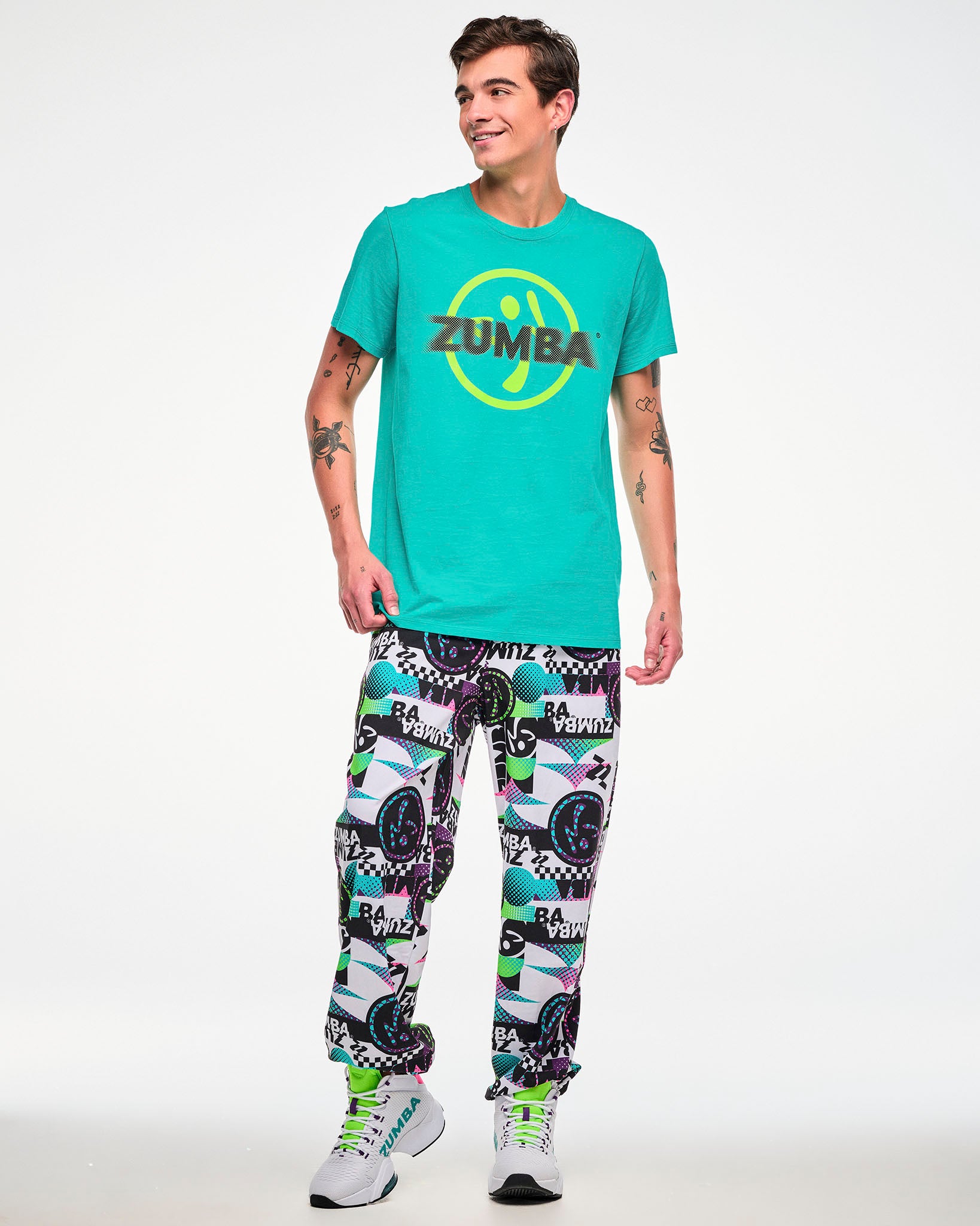 Male model wearing an aqua tee with a yellow Zumbito logo and black "ZUMBA" text, paired with white track pants featuring an all-over abstract print and white sneakers.