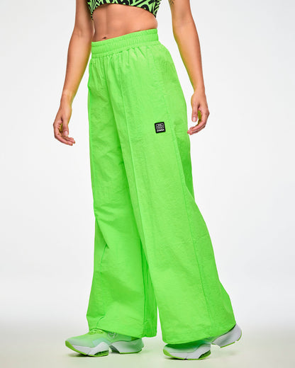 Female model wearing green wide-leg track pants with a cassette tag patch on the upper left leg and paired with matching green shoes.