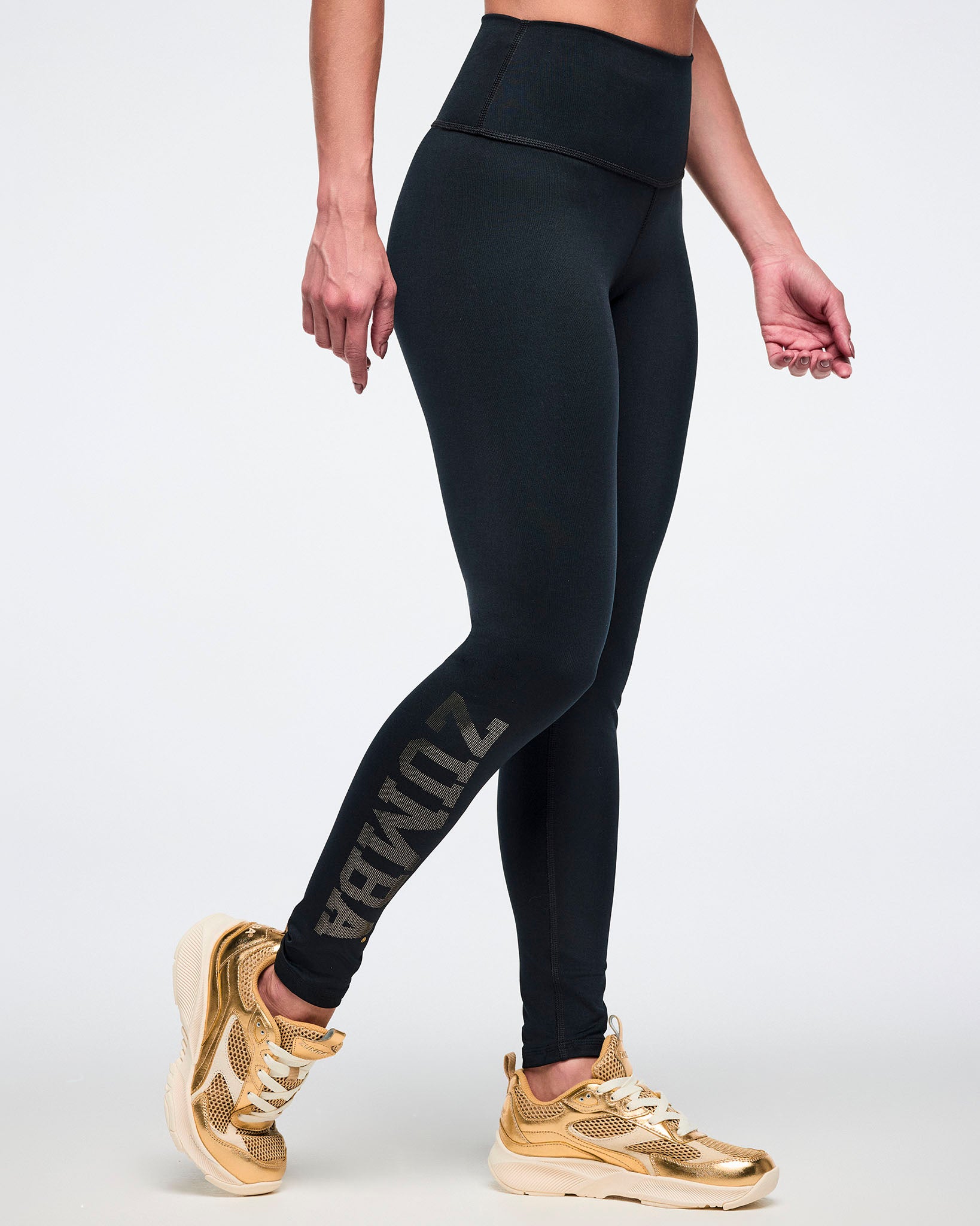 Female model wearing black leggings with gold text and gold sneakers on a white background.