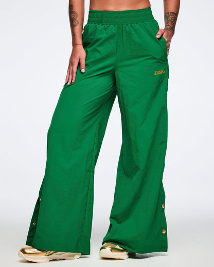 Female model on a white background, left hand in pocket, wearing green wide-leg pants with gold side snaps and gold shoes.