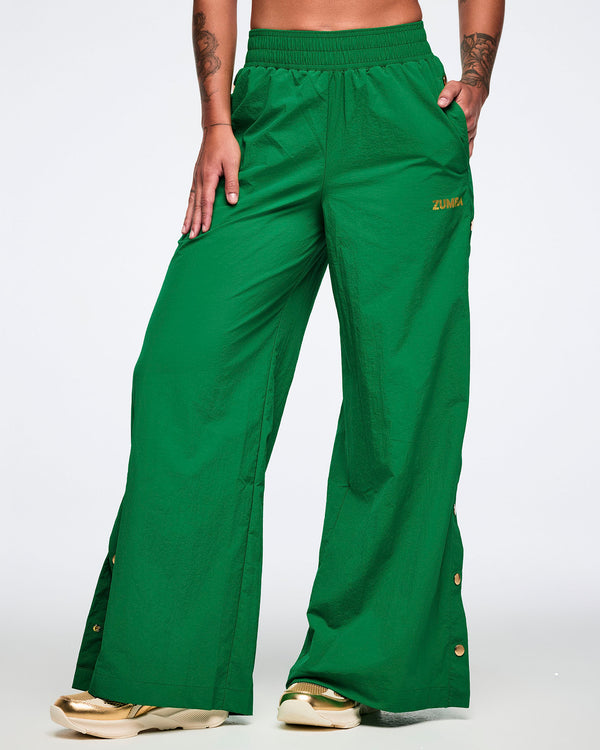 Female model on a white background, left hand in pocket, wearing green wide-leg pants with gold side snaps and gold shoes.