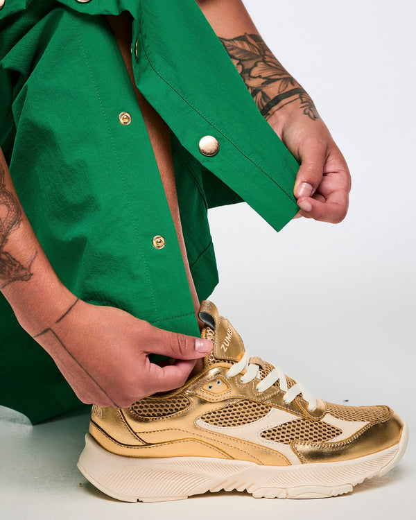 Female model on a white background, unsnapping green cargo pants with gold side snaps and wearing gold Zumba shoes.