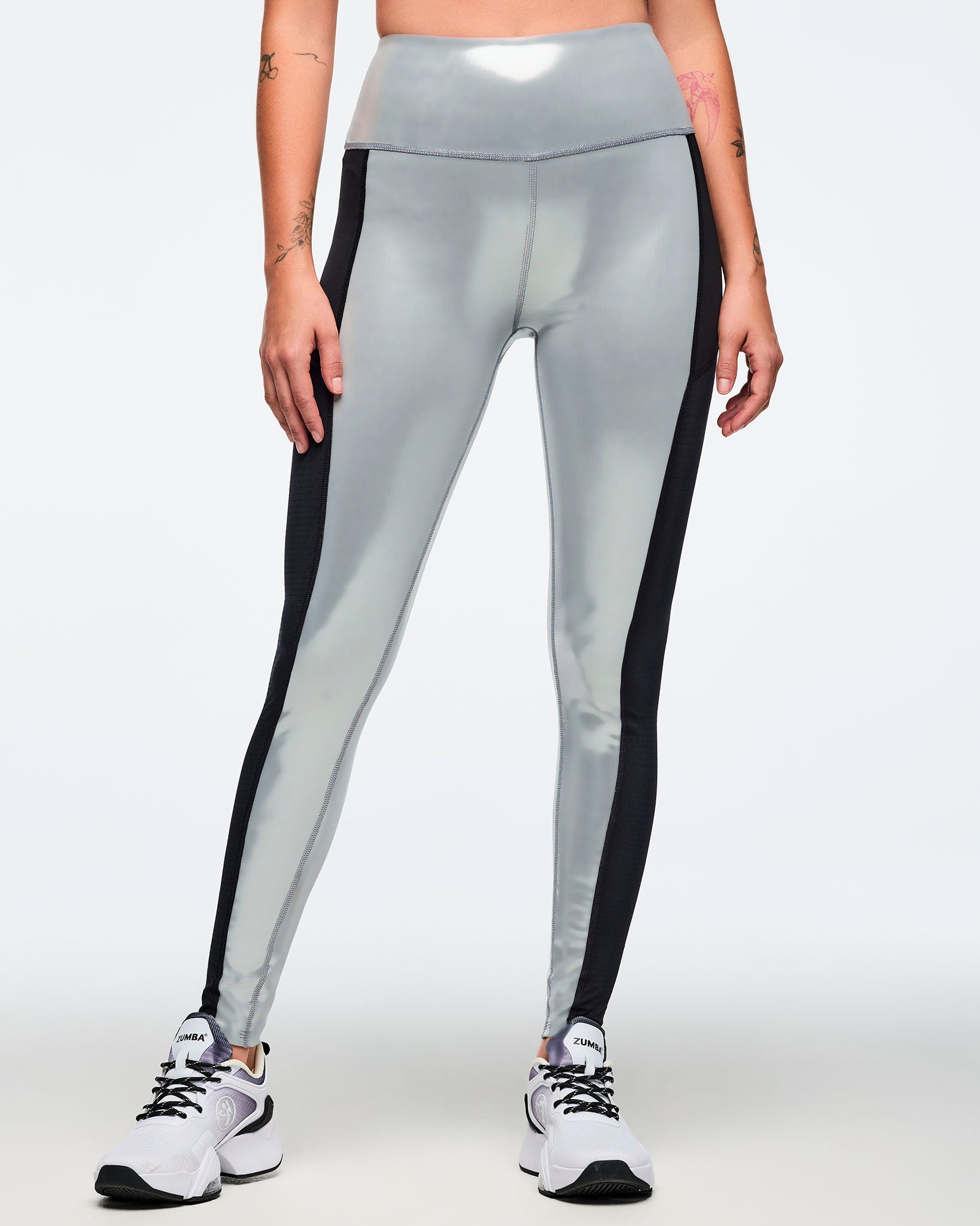Metallic silver high-waisted leggings with black side panels and a glossy finish. Female model wears fitted full-length leggings with a wide waistband, shown in a front close-up with white and black athletic sneakers against a light gray background.