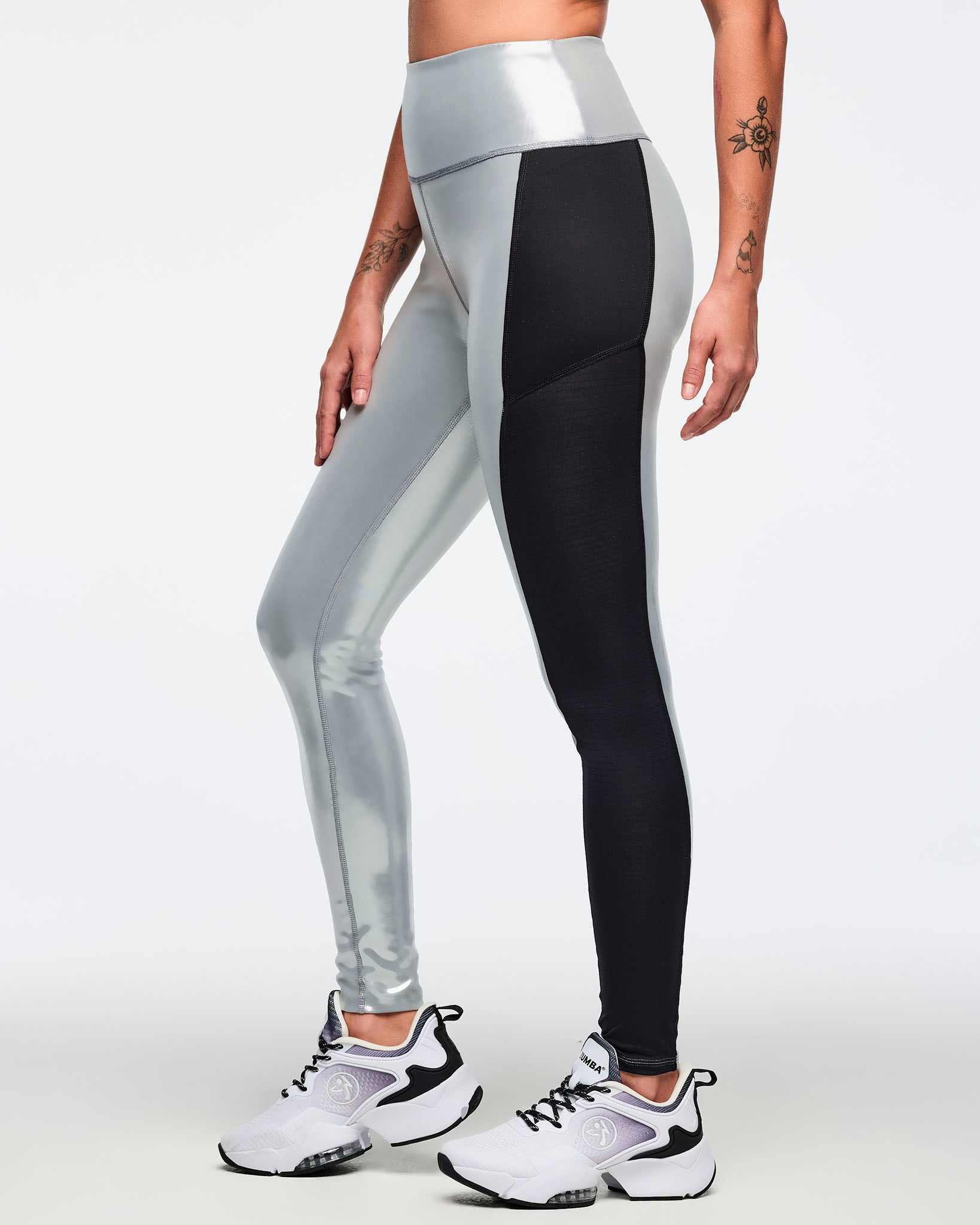Metallic silver high-waisted leggings with black side panels. Female model wears fitted glossy full-length leggings with a wide waistband and white athletic sneakers, shown in a side view against a light gray background.