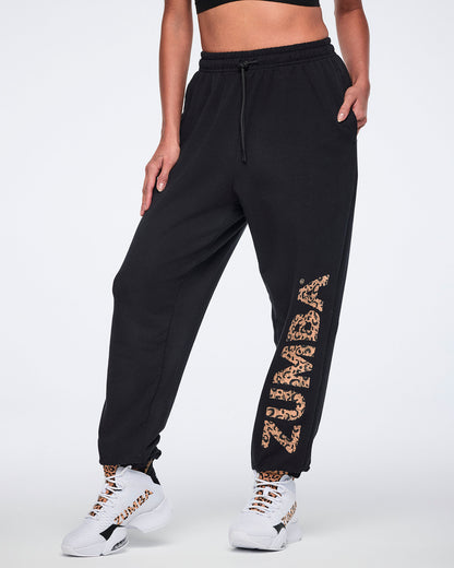 Angled front view of black sweatpants with an elastic drawstring waistband, tapered leg, side pockets, and leopard-print “ZUMBA” graphic down one leg. Female model wears the pants with white high-top Zumba sneakers.