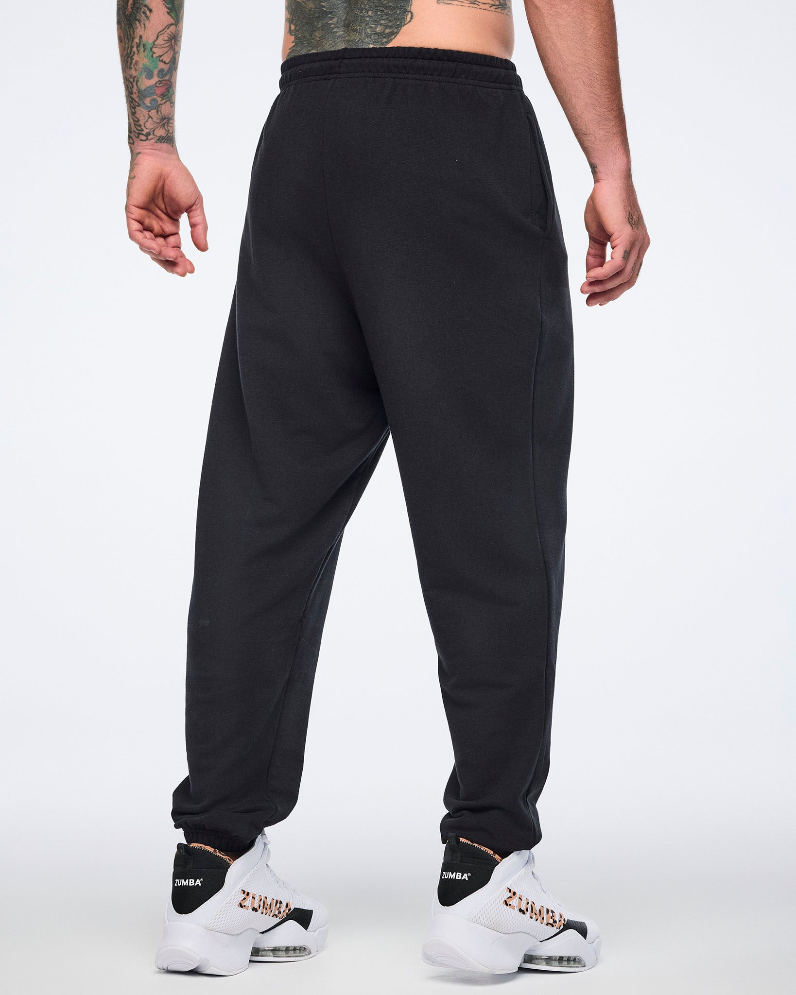 Back view of black sweatpants with an elastic waistband, tapered leg, and gathered cuffs. Male model wears the pants with white high-top Zumba sneakers.