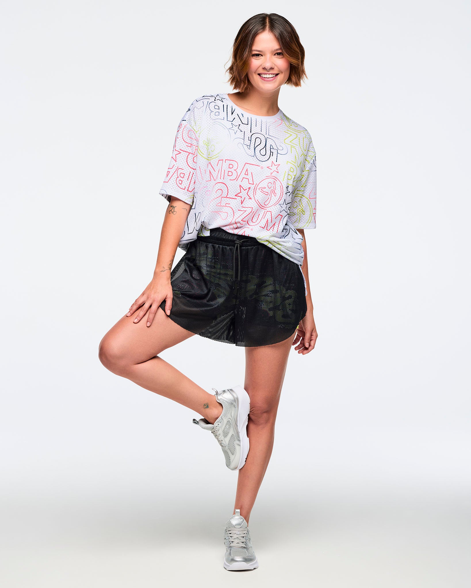 Female model wears a loose white oversized Zumba graphic T-shirt, sheer black mesh shorts with a drawstring elastic waistband and inner biker shorts, and silver athletic sneakers, posing against a light gray background.