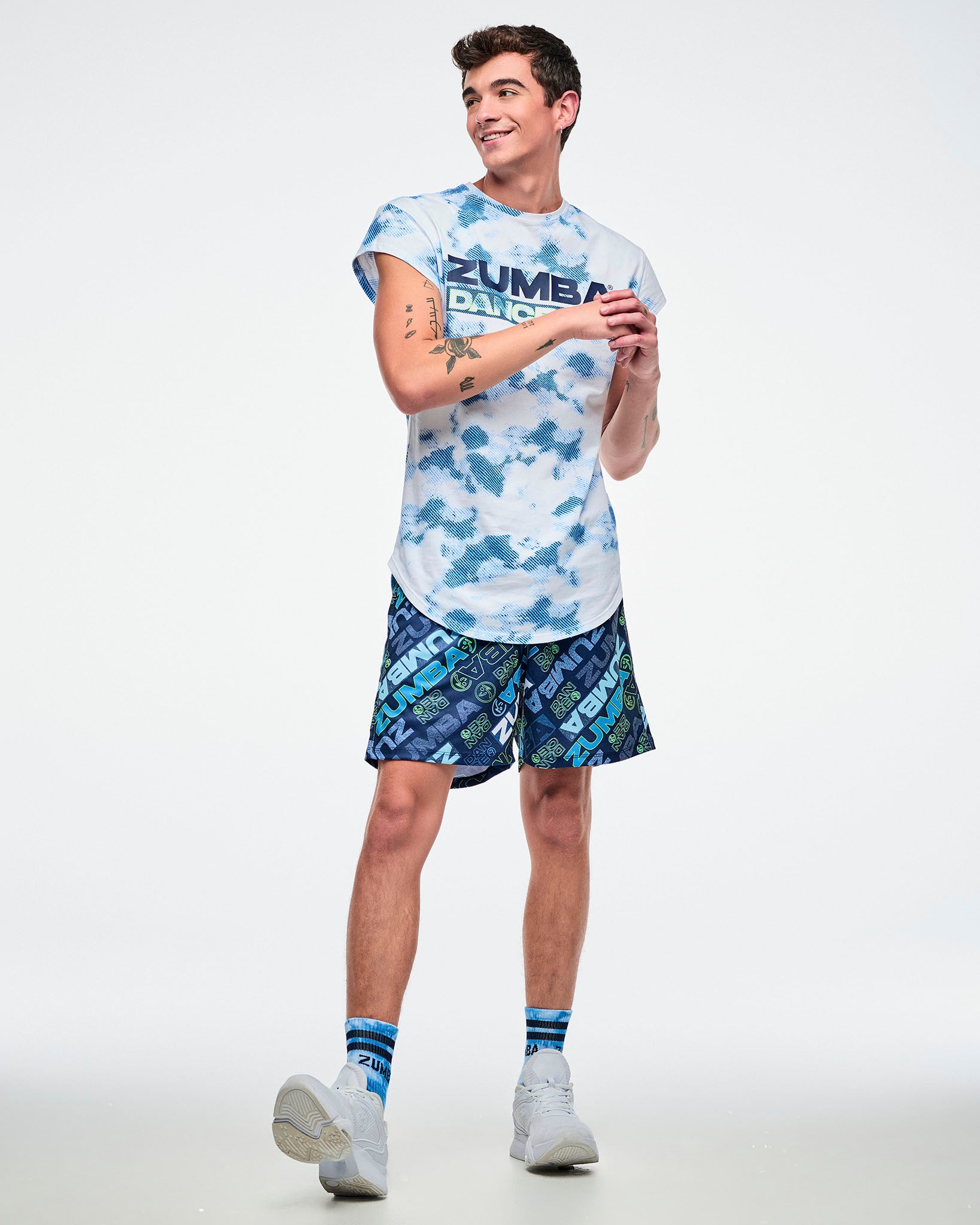 Full view of a male model in blue patterned shorts with both hands in his pockets, paired with a blue-and-white top and white shoes.