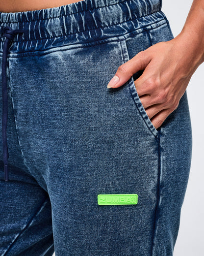 Close-up of a female model with her left hand in the pocket of blue denim pants featuring a green label.