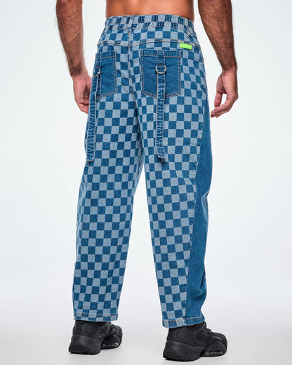 Back view of a male model in the checkerboard barrel ankle pants, showing an elastic back waistband, a lime green 'ZUMBA' patch, solid denim back pockets, and denim straps with silver D-rings hanging from each pocket.