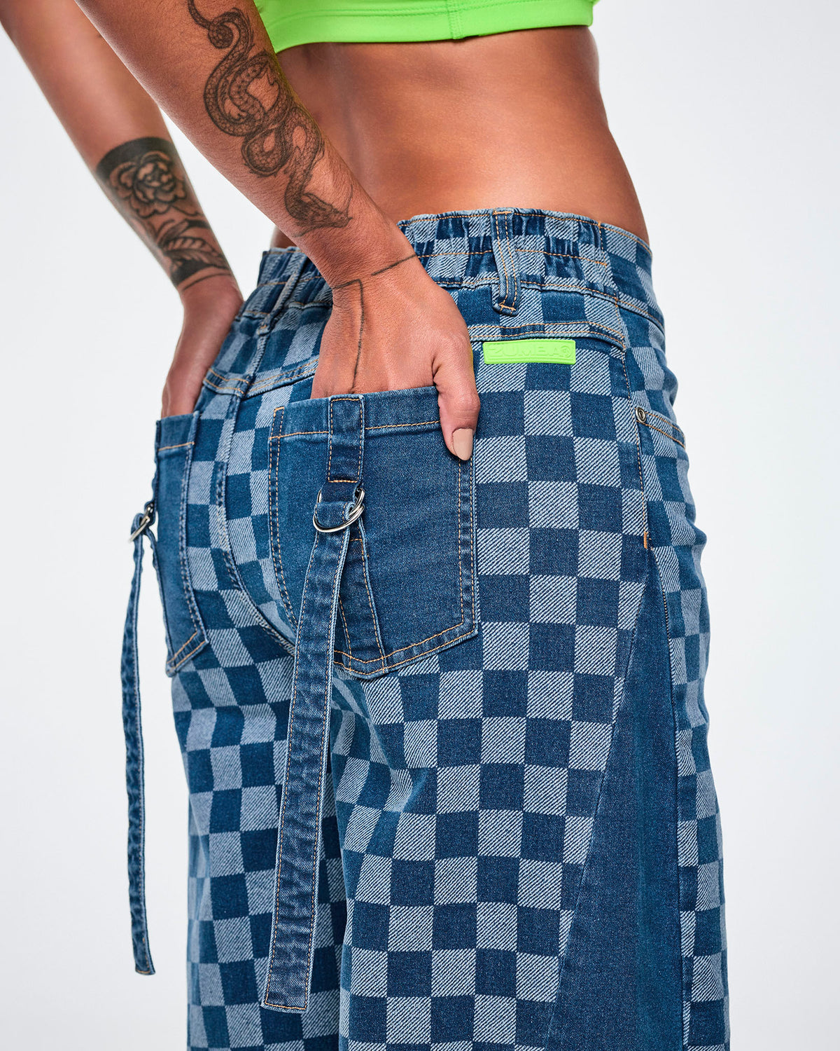 Close-up of the back of the barrel ankle pants, showing the lime green rubber 'ZUMBA' patch at the waistband and solid denim back pockets, with denim straps looped through silver D-rings.