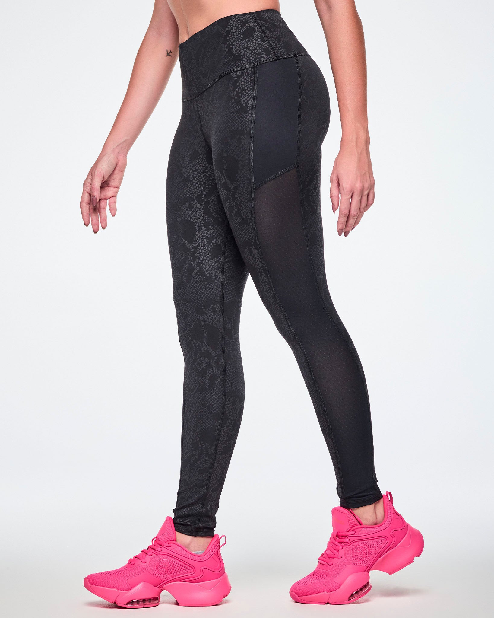 A female model wearing black high-waisted leggings with a subtle tonal snake-skin pattern and sheer mesh side panels, paired with pink shoes.
