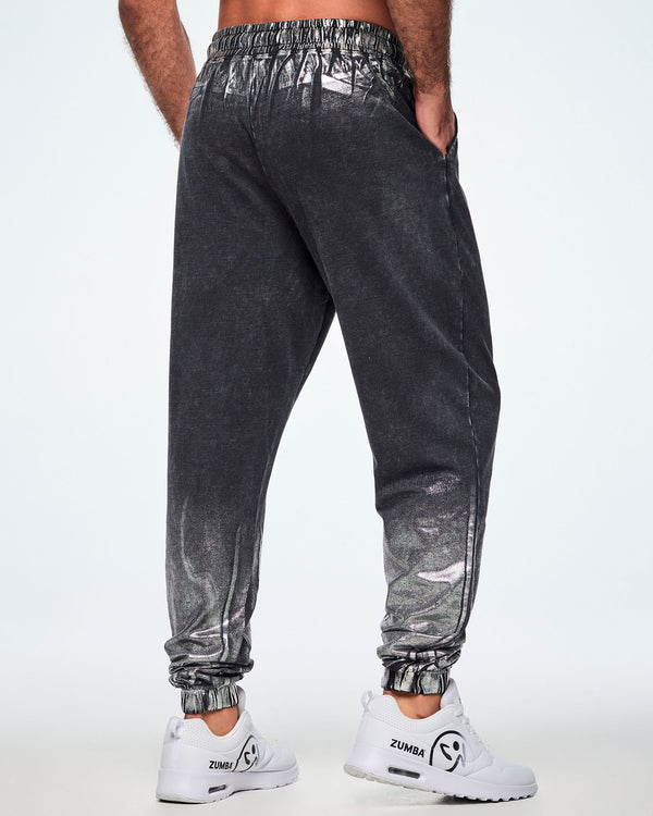 Back view of a male model wearing black and silver dark acid-wash jogger sweatpants with a subtle tonal pattern, featuring side pockets and elastic cuffs, paired with white shoes.