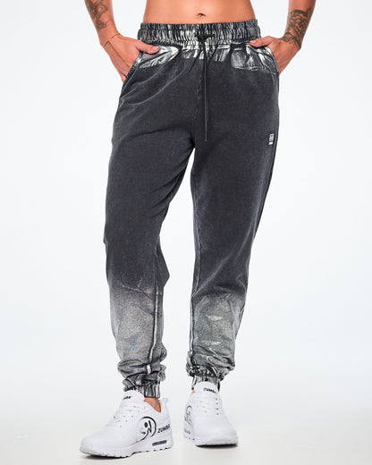 Front view of a female model wearing black and silver dark acid-wash sweatpants with a subtle tonal pattern, featuring a drawstring elastic waist, side pockets, and elastic cuffs, paired with white shoes.