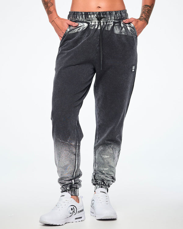 Front view of a female model wearing black and silver dark acid-wash sweatpants with a subtle tonal pattern, featuring a drawstring elastic waist, side pockets, and elastic cuffs, paired with white shoes.