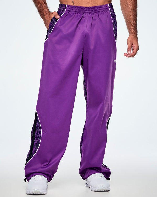 Front view of a male model, a hand in his pocket, wearing purple track pants with purple and black snake-skin patterned side panels, small white Zumba text on the left leg, white curved piping and paired with white shoes.