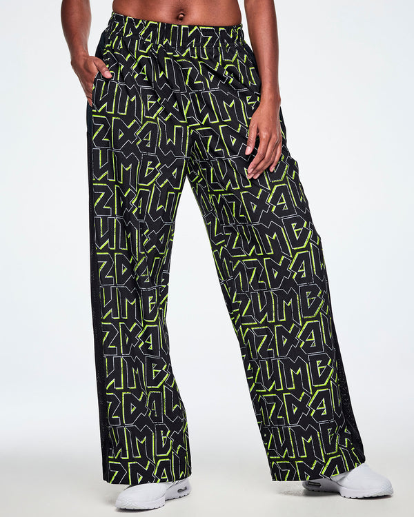 Front view of a female model wearing wide leg track pants, with ZUMBA text in an all over black pattern outlined in Zumba green colors ,with a black mesh side panel strip and paired with white shoes.