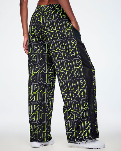 Back view of a female model wearing wide leg track pants, with ZUMBA text in an all over black pattern outlined in Zumba green colors with a black mesh side panel strip and paired with white shoes.
