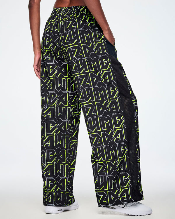 Back view of a female model wearing wide leg track pants, with ZUMBA text in an all over black pattern outlined in Zumba green colors with a black mesh side panel strip and paired with white shoes.