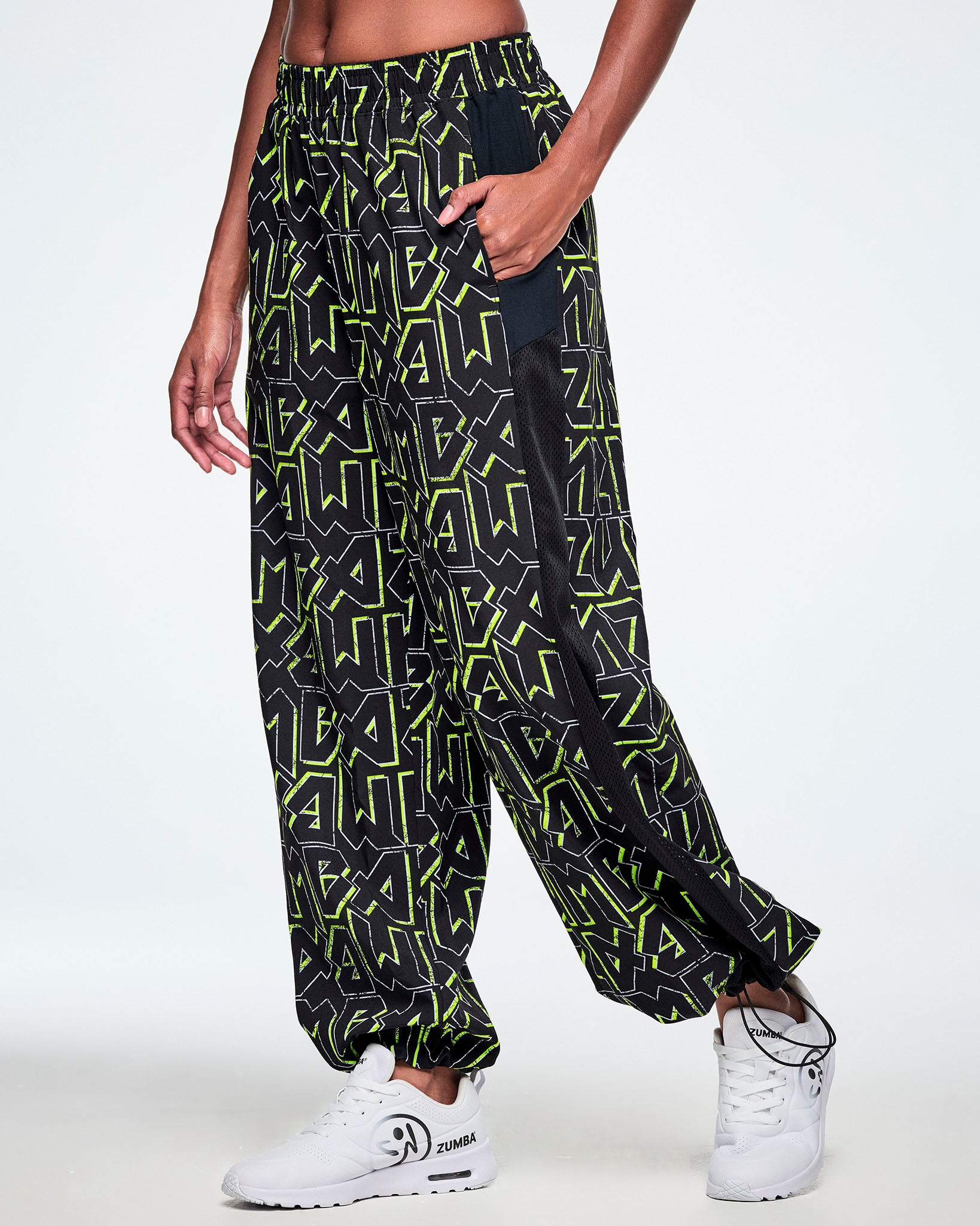 Side view of a female model with her hand in the side pocket of the wide leg track pants, with ZUMBA text in an all over black pattern outlined in Zumba green colors, with a black mesh side panel strip and paired with white shoes.